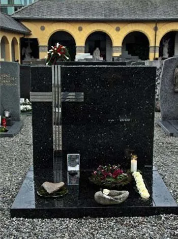 Schwarzes Grabmal mit hellem Kreuz und Glasstele vor einer Friedhofsarkade