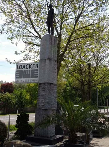 Hohe Stele mit Figur auf dem Ausstellungsgelände, daneben das Firmenschild